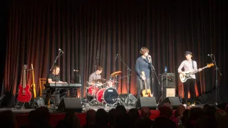 Eine Band spielt auf der Bühne mit einem roten Vorhang im Hintergrund. Ein Mann singt, während ein anderer Mann Gitarre spielt. Ein weiterer Mann spielt Schlagzeug. Es gibt einen Mann, der Klavier spielt. Auf der Bühne stehen Lautsprecher.
