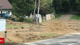 Eine schlammige Straße mit Schmutz und Trümmern, einem Pfosten in der Mitte und mehreren Bäumen und Sträuchern an der Seite. Es gibt auch einige Gegenstände wie Leitern und Planen.