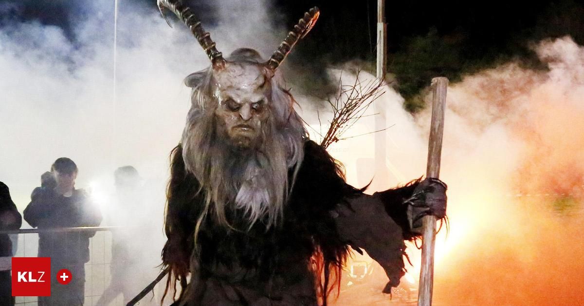 Eine Person in einem Kostüm mit Hörnern steht vor einem Feuer und hält einen Holzstab in beiden Händen. Das Kostüm ist schwarz und hat langes, weißes Haar und einen Bart.