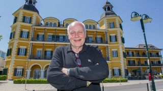 Ein lächelnder älterer Mann steht vor einem gelben Gebäude mit grünen Fensterläden, trägt ein schwarzes Hemd und eine Brille am Kragen.