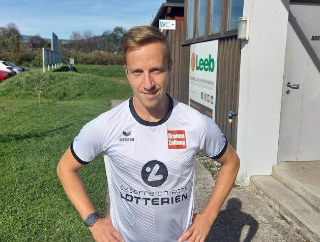 Ein Mann in einem weißen Shirt mit schwarzen Säumen posiert für ein Foto im Freien. Er steht vor einem Gebäude mit einem Schild, auf dem Leeb steht.