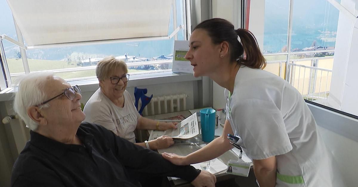 Eine lächelnde Frau liest eine Zeitung, während eine andere Frau und eine Krankenschwester neben ihr sitzen und alle auf die Krankenschwester schauen, die einen Stift hält. Hinter ihnen ist eine Aussicht auf das Meer und Gebäude.