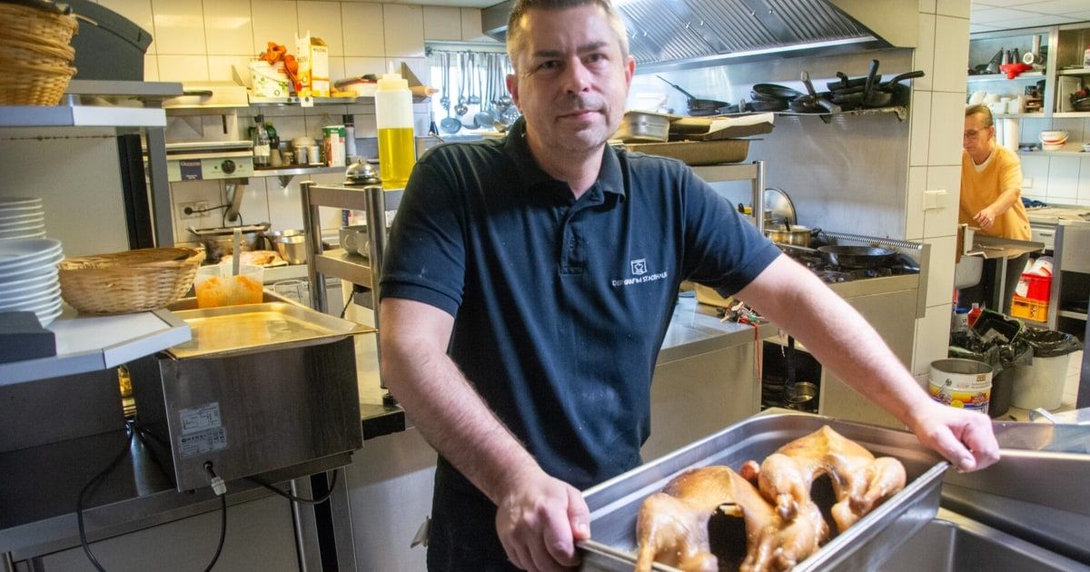 Ein Mann steht in einer Küche und hält eine Schale mit gekochten Vögeln, trägt ein blaues Hemd mit Logo. Hinter ihm sind Küchengeräte und Utensilien zu sehen.