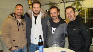 Vier Männer stehen nebeneinander und lächeln, tragen Winterjacken. Ein Mann hält eine Bierflasche. Es gibt einen Tisch mit Plastikbechern und einer kleinen Aschenschale.