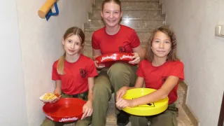 Drei junge Mädchen in roten Uniformen sitzen auf einer Treppe, lächeln und halten rote und gelbe Schüsseln, möglicherweise an einer Werbeveranstaltung teilnehmend.