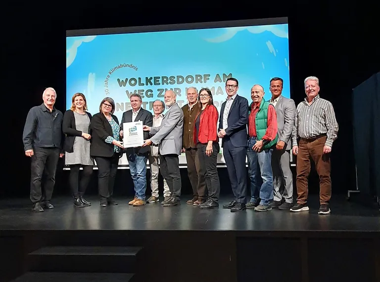 Eine Gruppe von Menschen steht auf einer Bühne und lächelt für ein Foto. Ein Mann in der Mitte hält ein Zertifikat und scheint es einer anderen Person zu geben. Hinter ihnen ist eine große Leinwand mit einem Text, der 'WOLKERSDORF AM WEG ZUR MA NET' lautet.