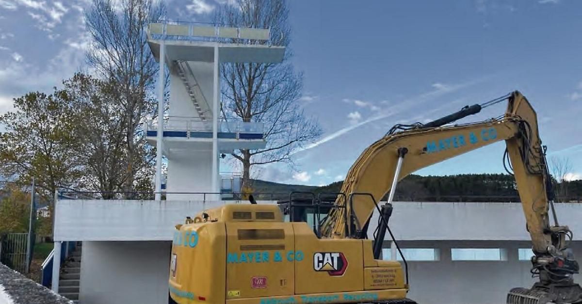 Ein gelber CAT-Bagger steht in der Nähe einer Sprungturmanlage, die eine weiße Plattform mit einem Metallzaun hat. Der Himmel ist klar mit einigen Wolken.