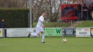 Ein Fußballspieler in weißer Uniform läuft auf einen Fußball auf einem Rasenfeld zu. Das Feld ist von einer Hecke und Bäumen umgeben. Banner mit Logos sind auf dem Zaun. Ein Mann beobachtet vom roten Kasten mit Autologo.
