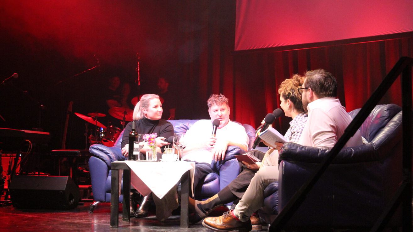 Vier Personen sitzen auf einer blauen Couch auf der Bühne, zwei halten Mikrofone, wahrscheinlich während einer Talkshow. Ein Tisch mit Weingläsern und einer Flasche steht zwischen ihnen. Rote Beleuchtung und Vorhänge sind im Hintergrund.