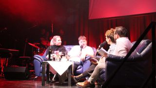 Vier Personen sitzen auf einer blauen Couch auf der Bühne, zwei halten Mikrofone, wahrscheinlich während einer Talkshow. Ein Tisch mit Weingläsern und einer Flasche steht zwischen ihnen. Rote Beleuchtung und Vorhänge sind im Hintergrund.