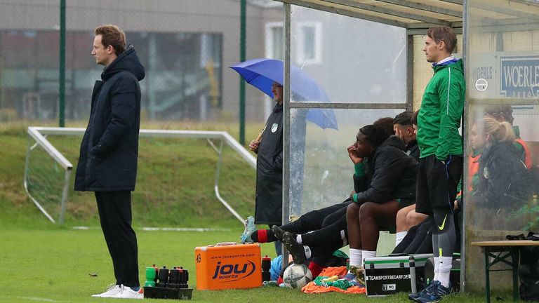 Ein Mann in einer schwarzen Jacke hält einen Regenschirm in der Nähe einer ruhenden Fußballmannschaft unter einem Schutzdach. Das Team trägt schwarze und grüne Trikots. Ausrüstung und ein Fußball liegen auf dem Rasen.