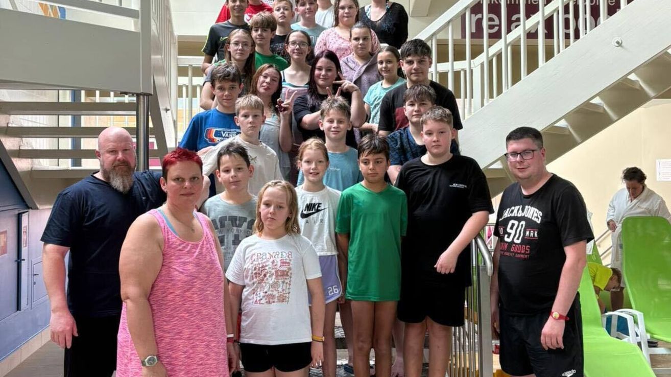 Eine Gruppe von Teenagern und zwei Erwachsene posiert für ein Foto auf einer Treppe, einige tragen Brillen und Turnschuhe. Ein Teenager trägt ein schwarzes T-Shirt, während andere Freizeitkleidung tragen.