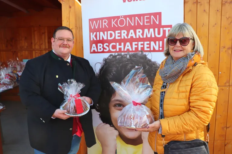 Two adults, a man and a woman, are standing next to each other in front of a poster, smiling. They are holding wrapped food items in plastic. The poster reads 'Wir konnen Kinderarmut abschaffen' in red and white.
