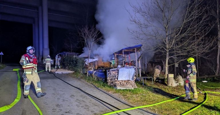 Firefighters extinguish a fire in a small shed surrounded by trees, with smoke billowing into the air. The scene is set under a bridge.