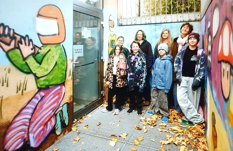 A group of people stand outside near a mural with autumn leaves on the ground. Some wear jackets, and two hold portraits. A woman in the center wears glasses.