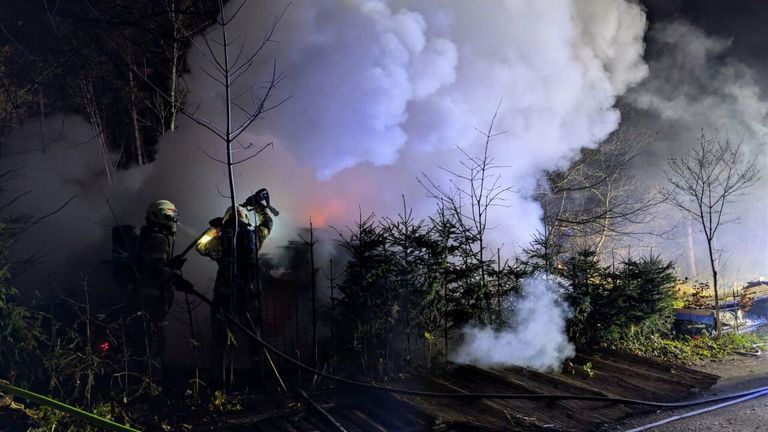 Firefighters are battling a blaze in a forest area, using hoses to spray water. Smoke fills the air, and a fire burns in the background.