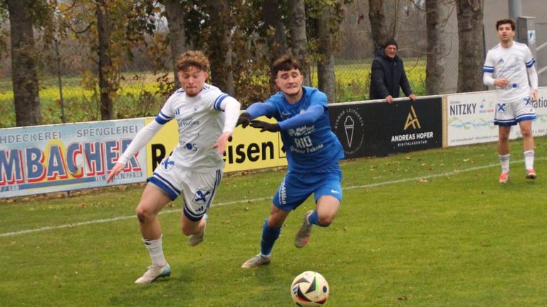 Two young soccer players are running on a grassy field. The player in blue is trying to kick the ball. Behind them, a man stands near a fence with advertisements.