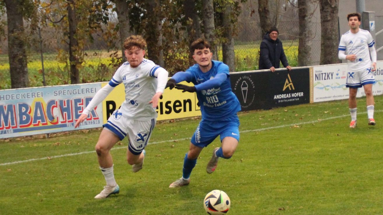 Two young soccer players are running on a grassy field. The player in blue is trying to kick the ball. Behind them, a man stands near a fence with advertisements.