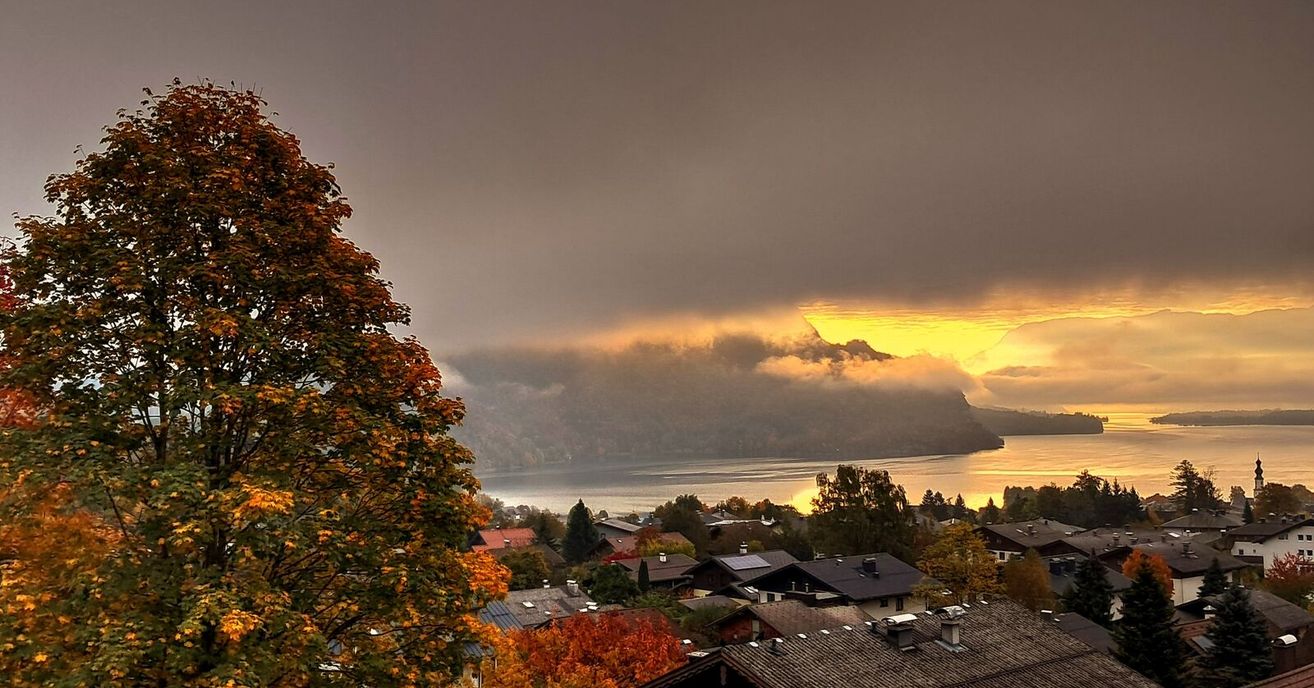 A village with houses, trees, and mountains. The sun is setting behind the clouds.