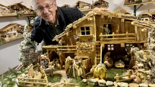 Ein älterer Mann steht neben einer detaillierten Krippe. Die Szene umfasst ein Holzhaus, Figuren von Jesus, Maria und verschiedenen Tieren, die in einer winterlichen Landschaft mit Felsen und Zweigen platziert sind.