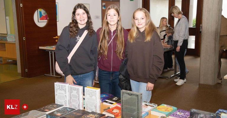 Three young girls stand next to each other in front of a book display, smiling for the camera. Behind them, a few people are standing, including a woman in glasses.