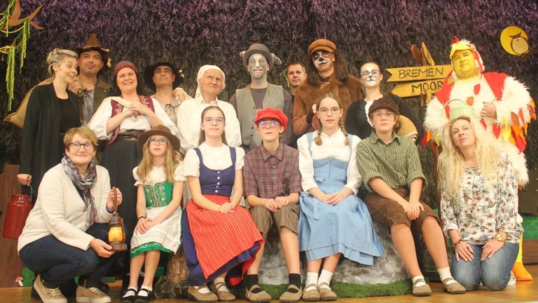 Eine Gruppe von Menschen in Kostümen posiert für ein Foto. Sie sitzen auf einem Felsen, und einige stehen hinter ihnen. Sie alle lächeln und schauen zur Kamera. Eine Lampe ist auf der linken Seite, und ein Schild ist auf der rechten Seite. Der Hintergrund ist unscharf.