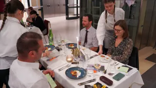 Eine Gruppe von Menschen sitzt an einem Tisch in einem Restaurant, bedient von einer jungen Frau mit geflochtenen Haaren. Der Tisch ist mit Tellern, Gläsern und Besteck gedeckt. Die Frau steht hinter dem Mann mit Bart. Auf dem Tisch stehen eine Kerze und eine Topfpflanze.