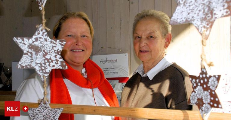 Zwei Frauen stehen in der Nähe einer Holzwand und lächeln für ein Foto. Eine trägt einen roten Schal und ein weißes Hemd, die andere ein braunes Sweatshirt und ein weißes Hemd. Ein dekorativer Stern ist im Vordergrund, und ein Schild lautet 'Kunsthandwerk'.