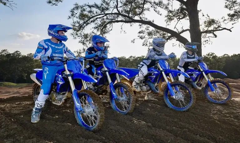 Four individuals in Yamaha jerseys and helmets pose with blue dirt bikes on a muddy track, with a tree in the background.