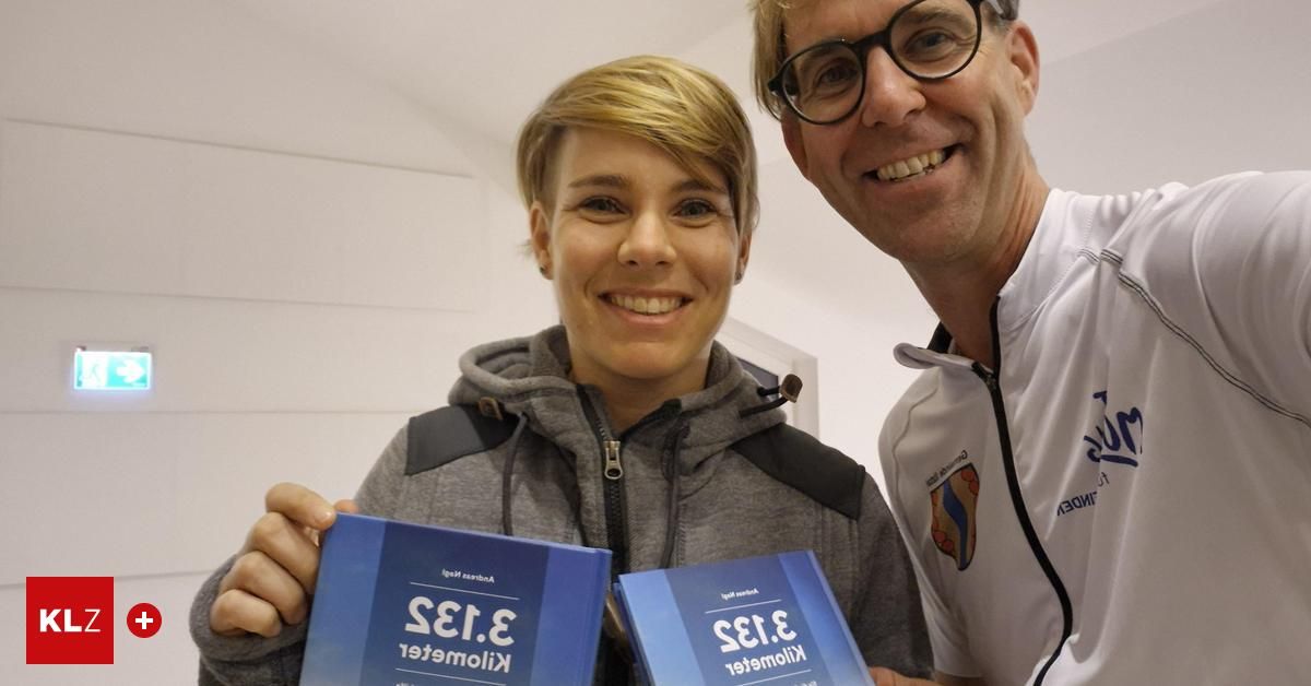 A smiling man and woman are standing together. The woman is holding two blue books with white text. They are posing for a photo.