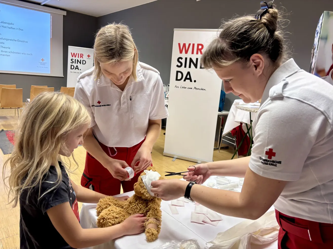 Drei Frauen verbinden einen braunen Plüschbären mit Gaze. Sie tragen rote Hosen und weiße Hemden. Die Frau auf der rechten Seite hält eine Schere und konzentriert sich auf den Bären. Hinter ihnen steht ein großes Schild, das 'Wir sind da' sagt.