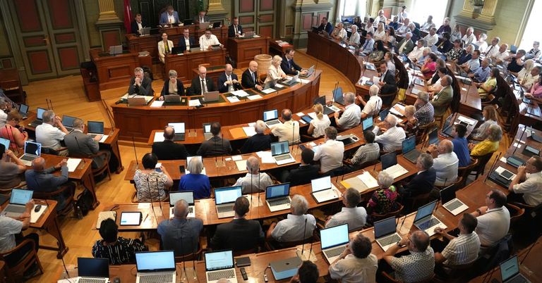 A large room with wooden tables and chairs is filled with people, many of whom are working on laptops. Some individuals are wearing ties, and others are dressed casually. The setting appears to be a conference or meeting hall with a flag visible in the background.
