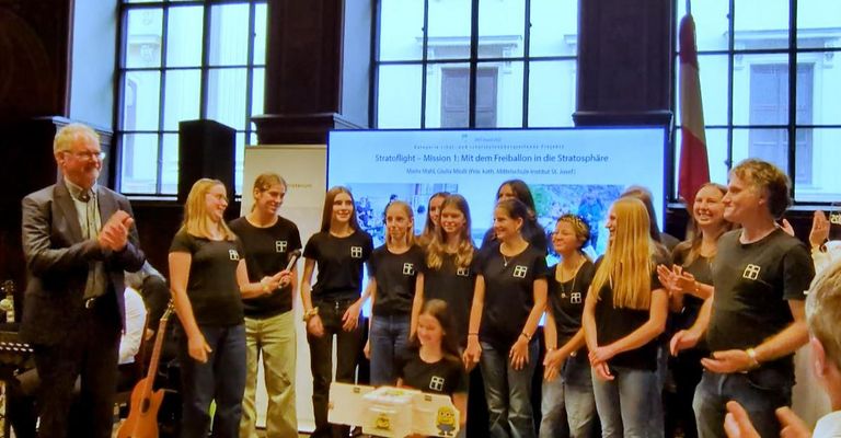 A group of people are standing in a room. They are wearing black T-shirts with logos. A person is holding a microphone. A girl is holding a box. A projector screen is mounted on the wall. A few people are standing in front of it. A speaker is placed on the left side.