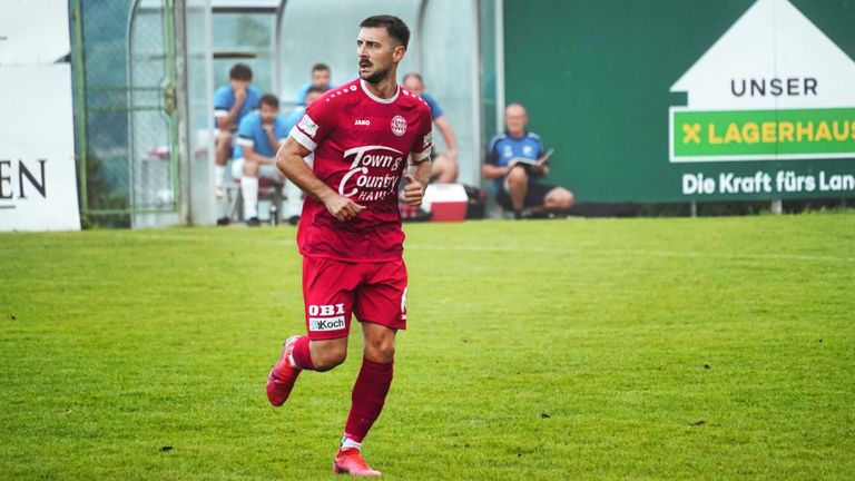 A soccer player in a red uniform with Town & Country written on it runs across the field. Behind him are teammates sitting on a bench. Another man sits on the ground reading a book.