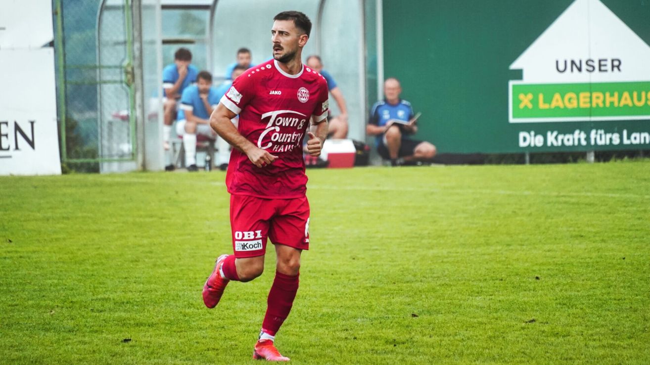 A soccer player in a red uniform with Town & Country written on it runs across the field. Behind him are teammates sitting on a bench. Another man sits on the ground reading a book.