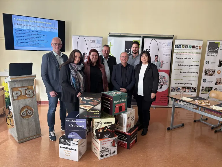 Mehrere Personen stehen um einen Stapel Kartons in einem Klassenzimmer und lächeln für ein Foto. Hinter ihnen zeigt ein Banner verschiedene Symbole und Text.