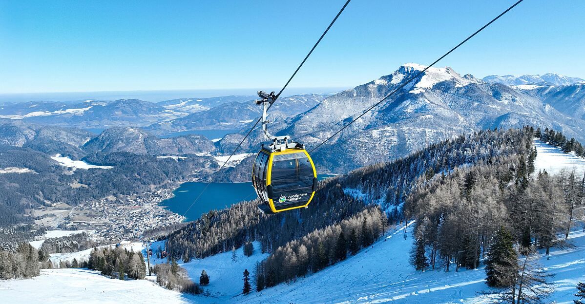 A cable car suspended over a snowy landscape, with a lake and snowy mountains in the distance.