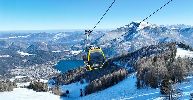 A cable car suspended over a snowy landscape, with a lake and snowy mountains in the distance.