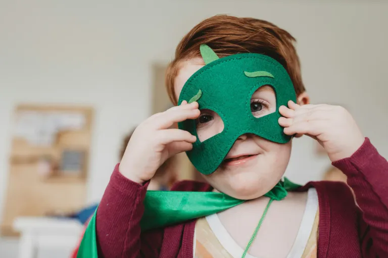 A young boy wearing a green mask with a cape smiles for the camera. He holds the mask up to his eyes.