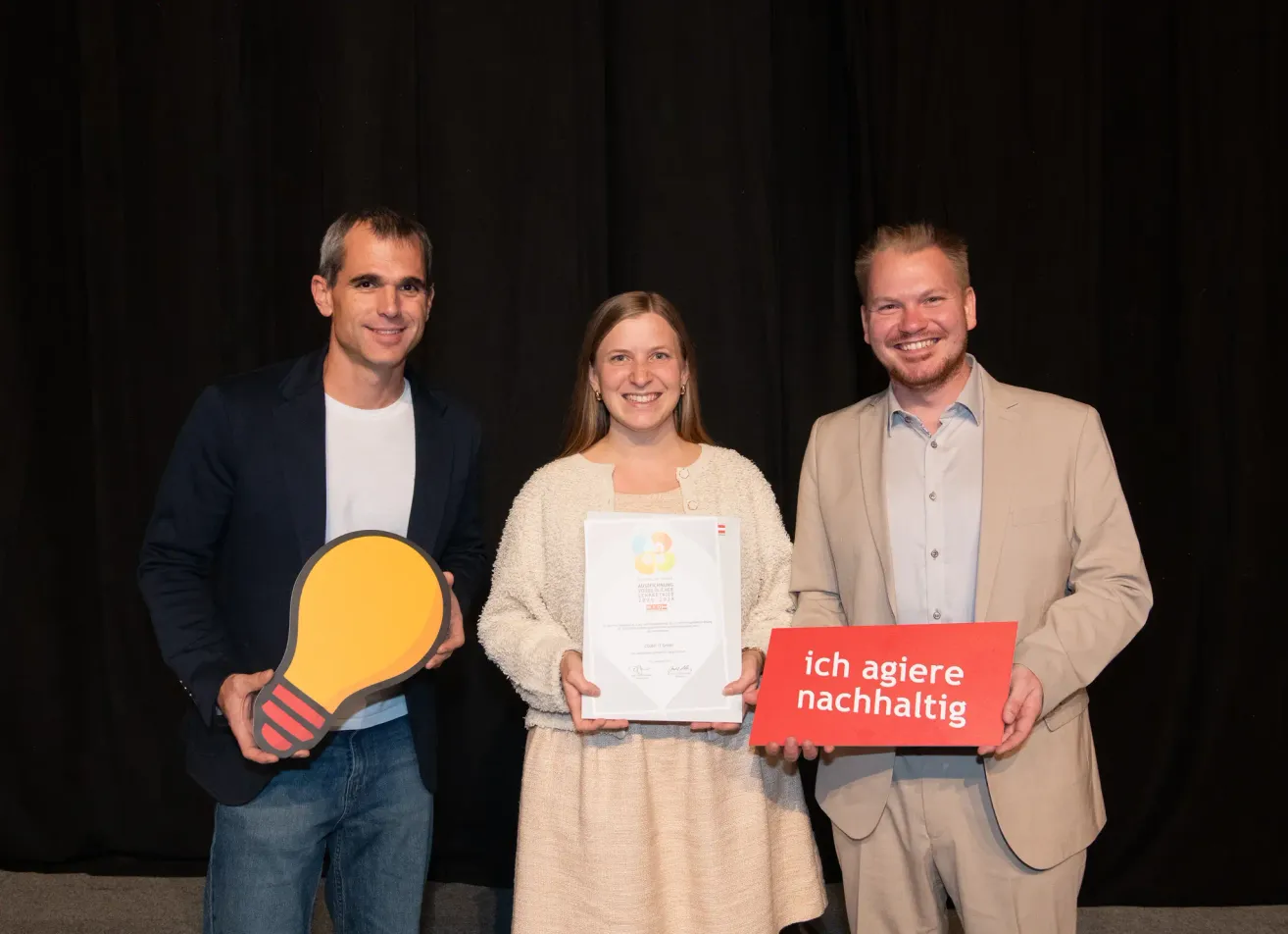 Three people stand in front of a black curtain, smiling. A man holds a lightbulb, a woman holds a certificate, and another man holds a sign.