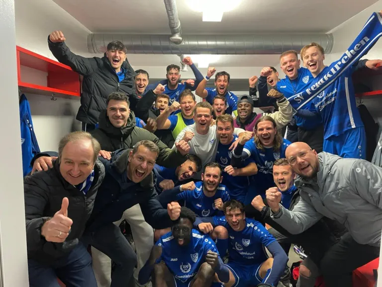 A group of soccer players and coaches in blue uniforms celebrate inside a locker room.