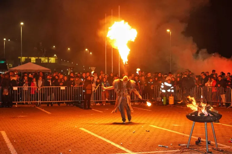 Ein Mann führt bei Nacht ein Feuershow vor einer Menge auf und hält einen Feuerspucker und eine Flasche in den Händen.