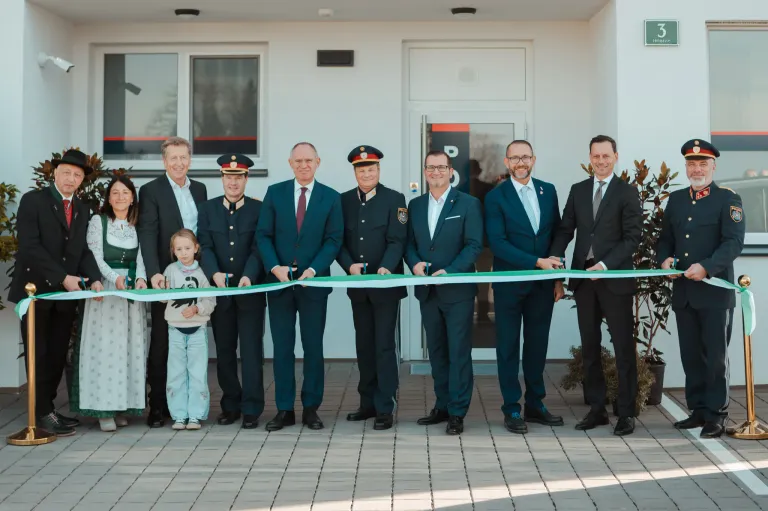 A group of people, including a child, are smiling and posing for a photo. They are standing in front of a building and holding a green ribbon, possibly for an opening ceremony.