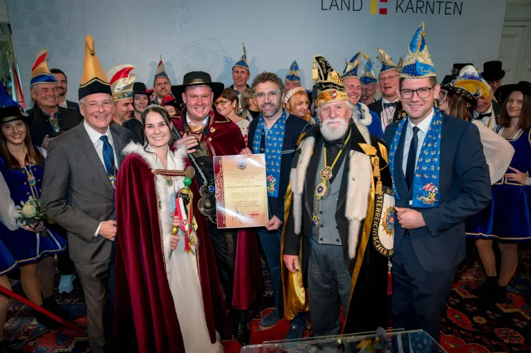 Group of people in traditional costumes, including a woman holding a certificate, pose for a photo in front of a wall with 'LAND KARNT' branding.