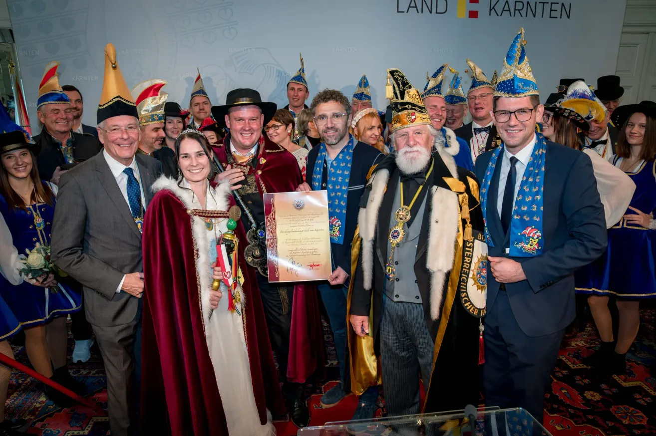 Group of people in traditional costumes, including a woman holding a certificate, pose for a photo in front of a wall with 'LAND KARNT' branding.