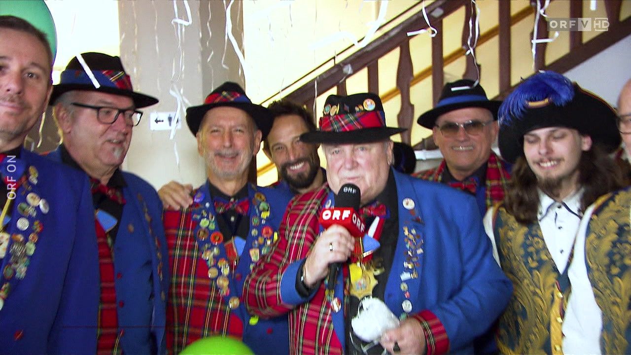 A group of men in matching blue and red tartan outfits, one holding a microphone and a toy dog, another wearing a red bow tie. They are smiling and appear to be posing for a photo.