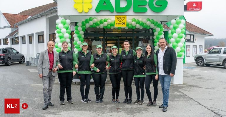 Eine Gruppe von Mitarbeitern in grünen und schwarzen Uniformen steht vor einem Laden mit einem grün-weißen Luftballonbogen. Der Laden hat ein Schild mit der Aufschrift ADEG bank 99.