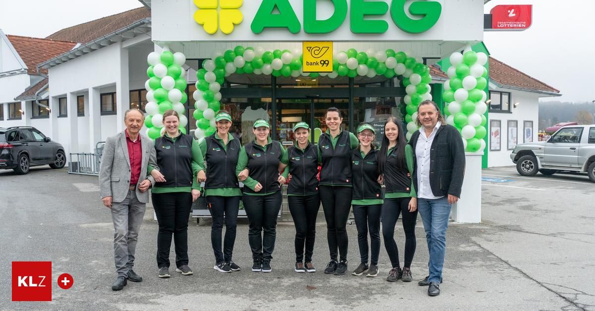 Eine Gruppe von Mitarbeitern in grünen und schwarzen Uniformen steht vor einem Laden mit einem grün-weißen Luftballonbogen. Der Laden hat ein Schild mit der Aufschrift ADEG bank 99.