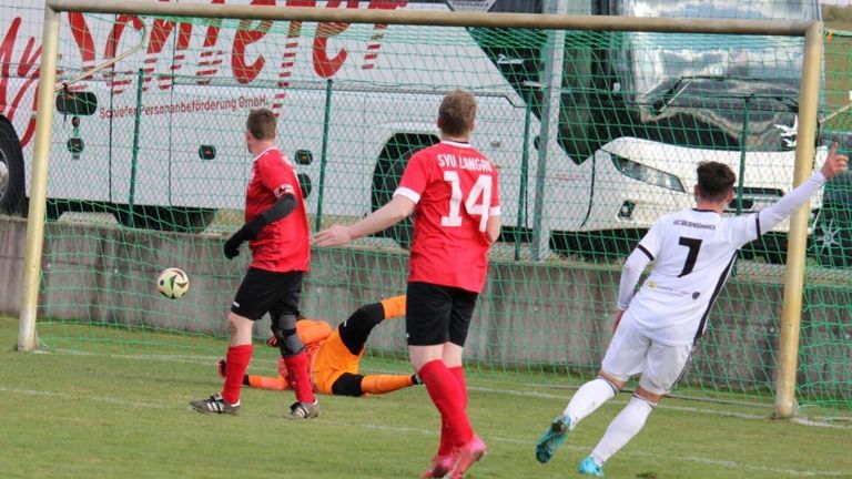 Drei Fußballer auf einem Spielfeld mit einem Bus und einem Zaun im Hintergrund. Ein Spieler in Rot geht, während zwei andere auf dem Boden liegen.
