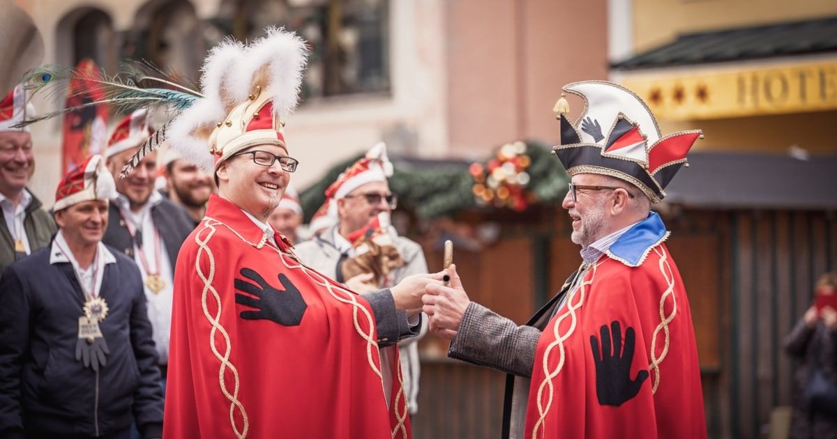 Two men in festive red and gold costumes and hats with feathers interact in a lively street setting, with others dressed similarly in the background.
